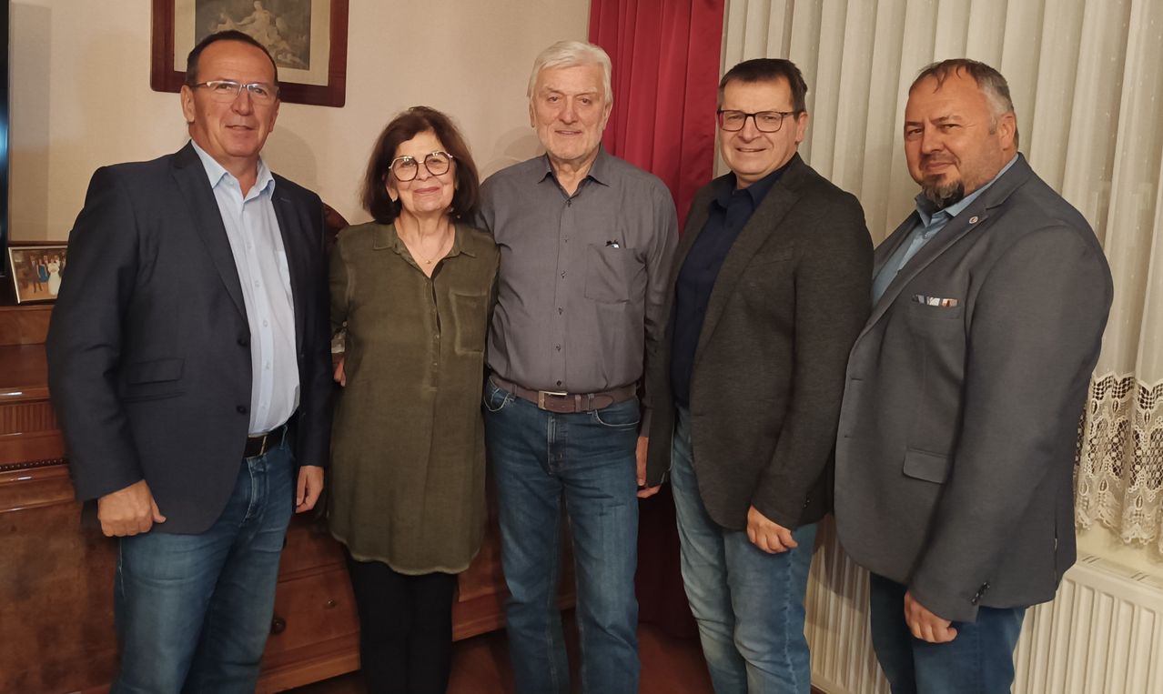 Fünf Personen posieren für ein Foto in einem Raum mit Holzschränken, einem roten Vorhang und einem Bilderrahmen an der Wand.