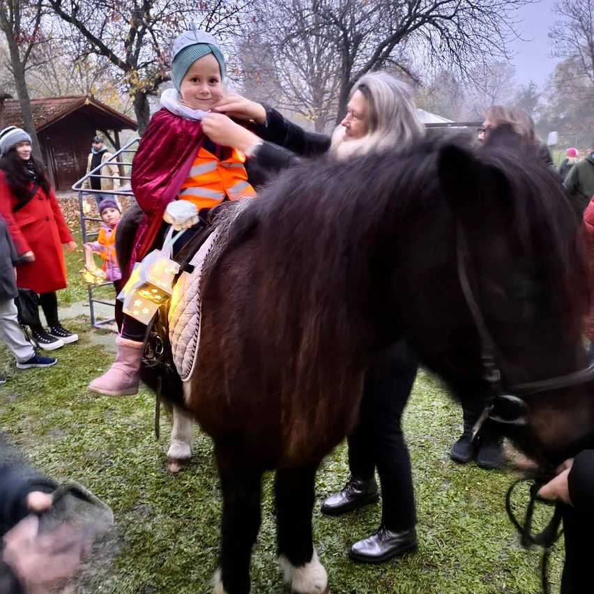 Eine Frau hilft einem Kind in einem roten Umhang, ein Pferd zu reiten. Sie haben Laternen an ihren Stiefeln. Eine kleine Gruppe von Menschen steht herum, einige in Winterkleidung.