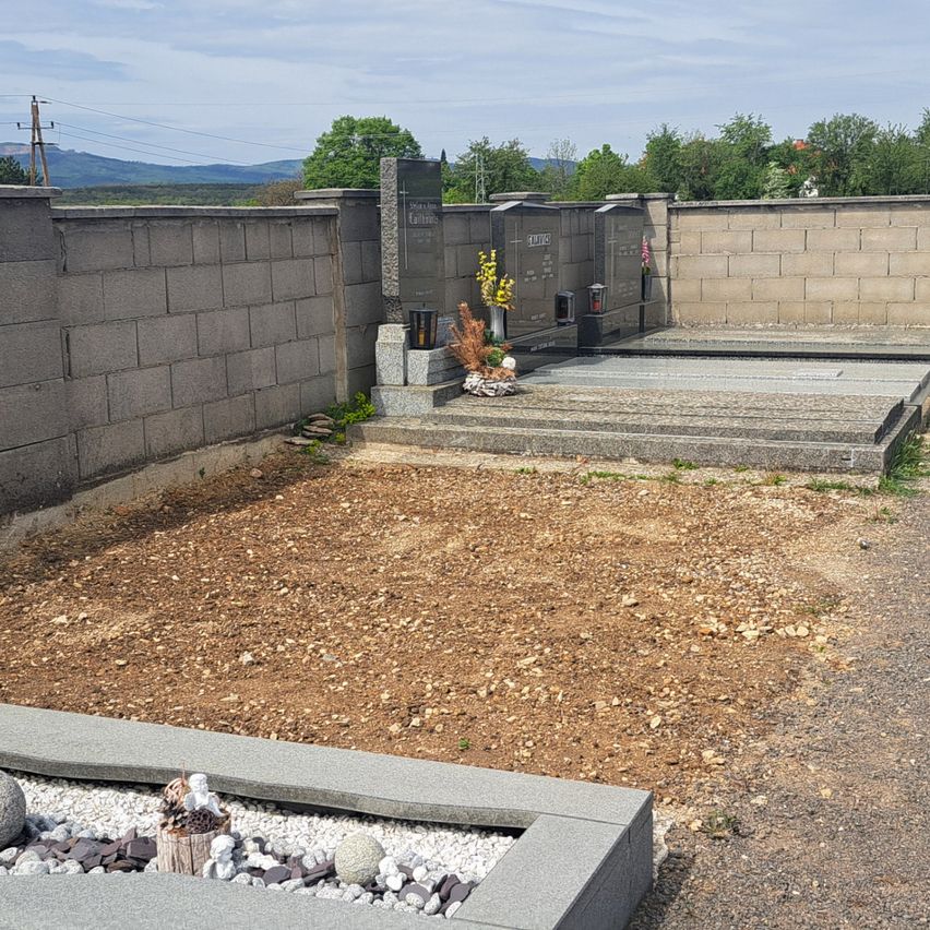 Ein Friedhof mit mehreren Grabsteinen, einige mit Blumen, vor einer Kulisse aus Bergen und Grün.