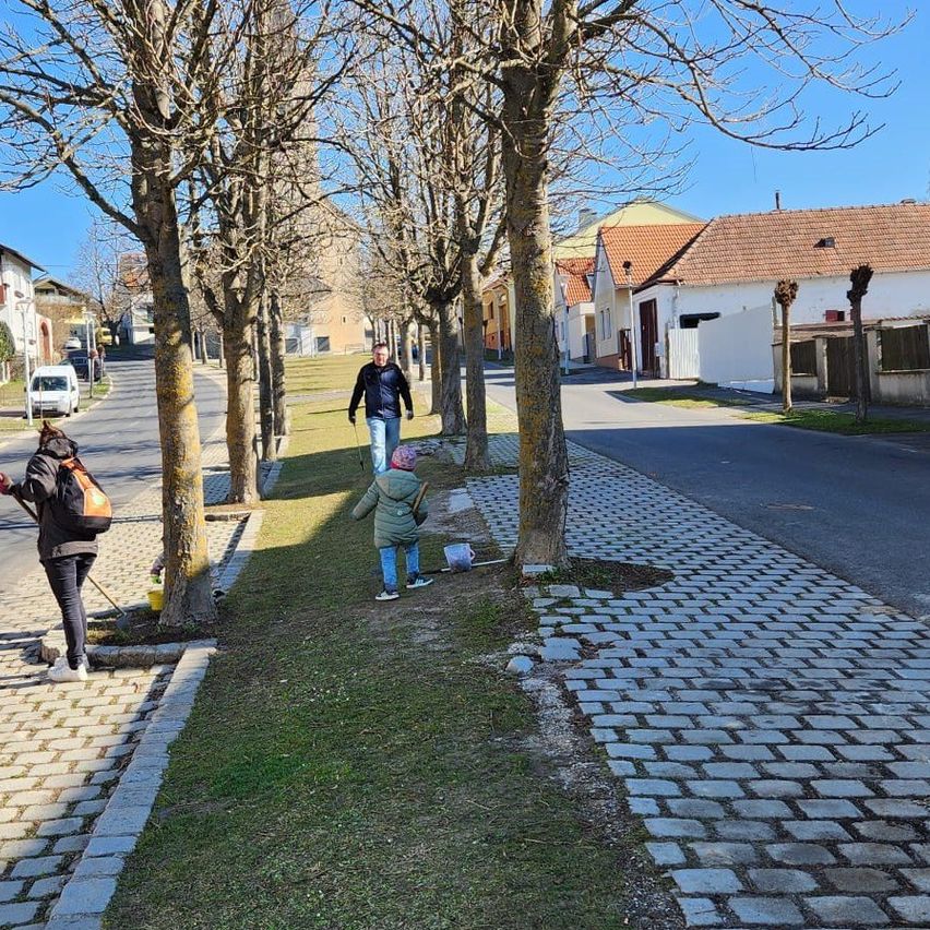 Menschen gehen auf einem Gehweg, gesäumt von Bäumen und einem gepflasterten Pfad, ein Kind hebt etwas auf. Im Hintergrund befinden sich Häuser mit Ziegeldächern und ein klarer Himmel.