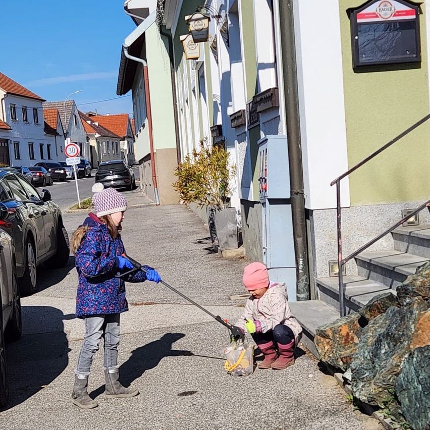 Zwei kleine Mädchen putzen die Straße. Eine hält einen Besen und die andere einen Beutel. Häuser und Autos sind am Straßenrand geparkt.