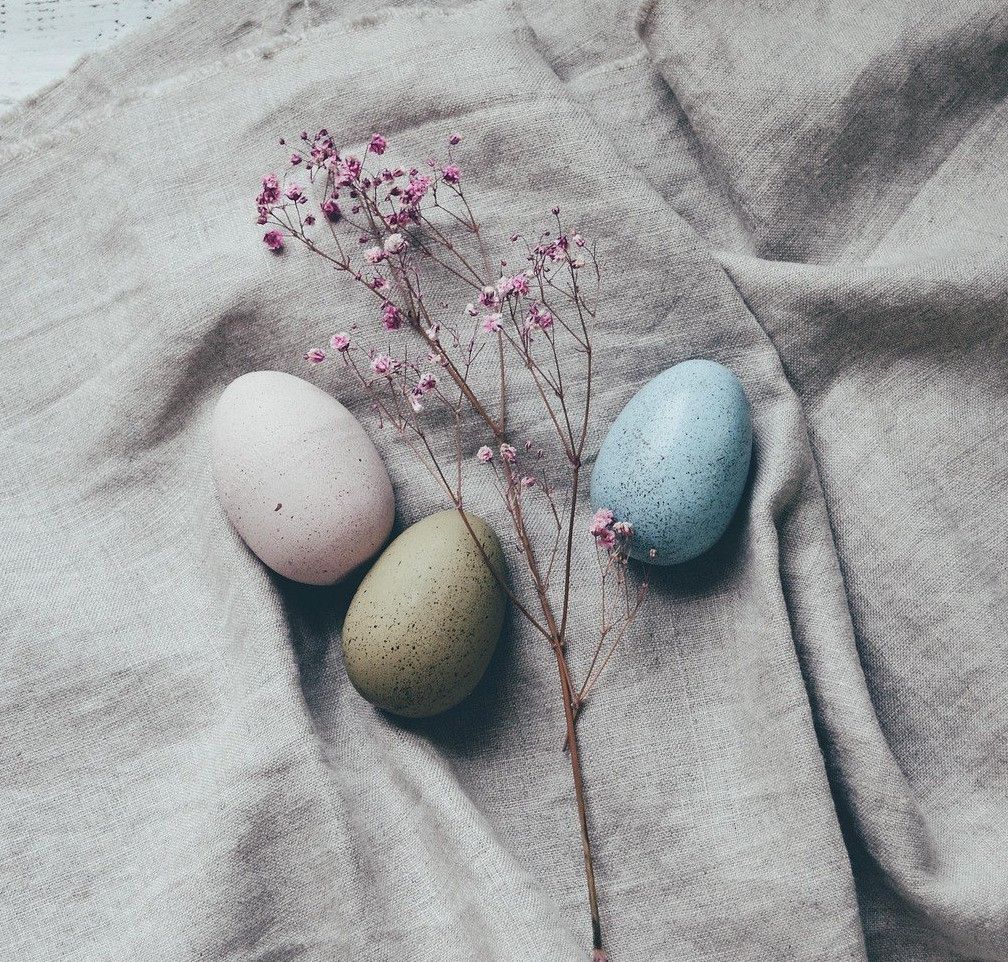 Drei Eier, ein rosa, ein blaues und ein geflecktes, liegen auf einem grauen Tuch mit kleinen rosa Blüten.