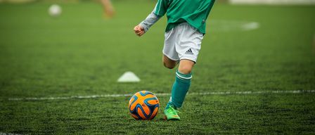 Ein junger Junge in einem grünen Fußballtrikot tritt einen Fußball auf einem Feld. Eine verschwommene Figur im Hintergrund läuft.