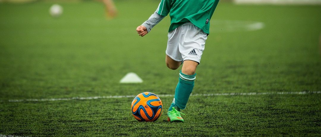 Ein junger Junge in einem grünen Fußballtrikot tritt einen Fußball auf einem Feld. Eine verschwommene Figur im Hintergrund läuft.