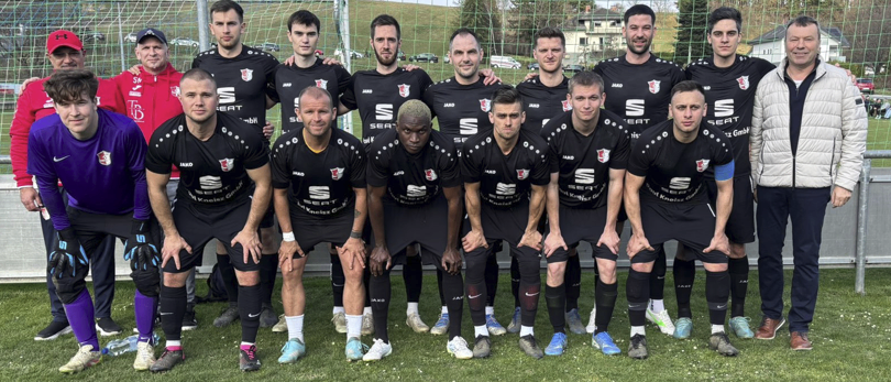 A soccer team, dressed in black, poses for a group photo on the field. Some wear gloves and sneakers, while two in the front wear hats. Behind them, a goalpost stands.