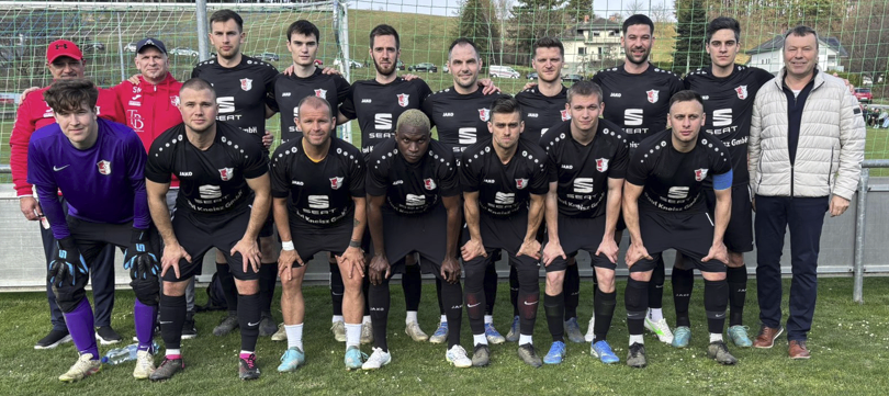 A group of soccer players in black uniforms pose for a team photo on a field. The players wear different numbers on their jerseys. They stand in front of a goalpost.
