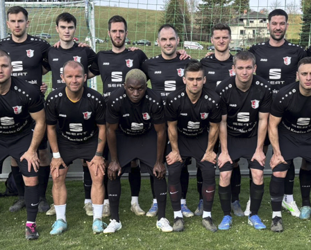 A group of soccer players in black uniforms pose for a team photo on a field. The players wear different numbers on their jerseys. They stand in front of a goalpost.