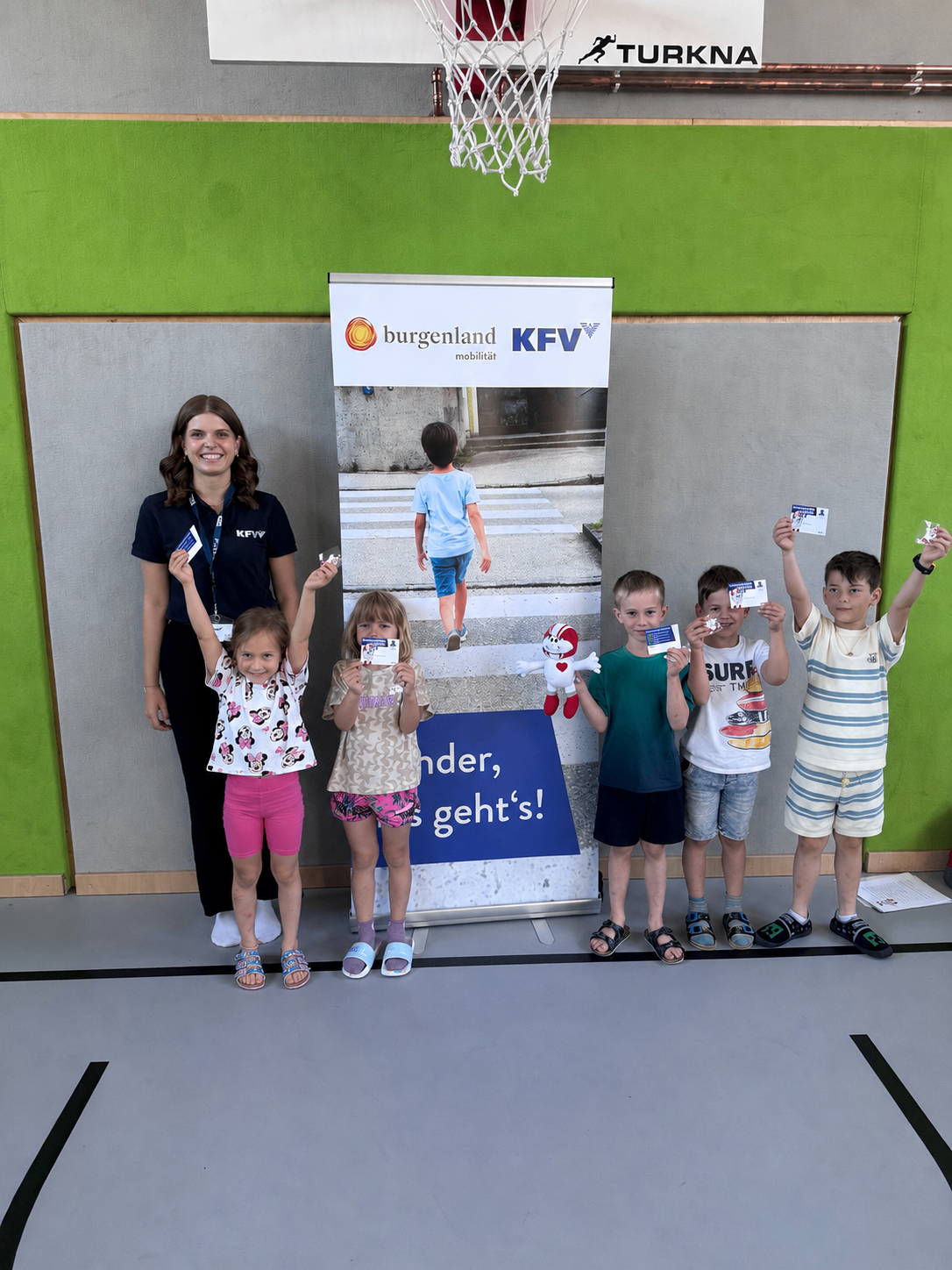 Eine Gruppe von Kindern und einem Erwachsenen steht vor einem Schild, das 'unter, geht's!' sagt. Die Kinder halten Karten hoch und lächeln. Eine Frau hält ein Kind. Der Raum hat grüne Wände und eine weiße Decke.