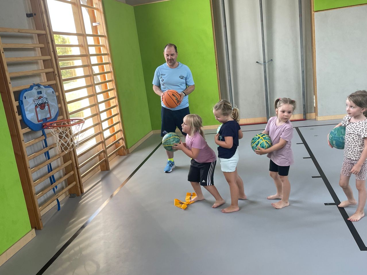 Ein Mann instruiert vier Mädchen in einer Turnhalle, jede hält einen Basketball. Die Wände sind grün, und in der Nähe befindet sich ein Basketballkorb.