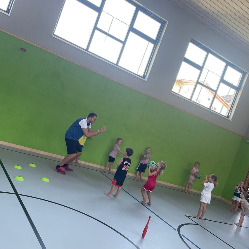 Eine Gruppe von Kindern und ein Mann spielen auf einem Indoor-Spielfeld mit grünen Wänden und Fenstern. Die Kinder halten Bälle und stehen in einer Reihe. Der Mann gibt Anweisungen.