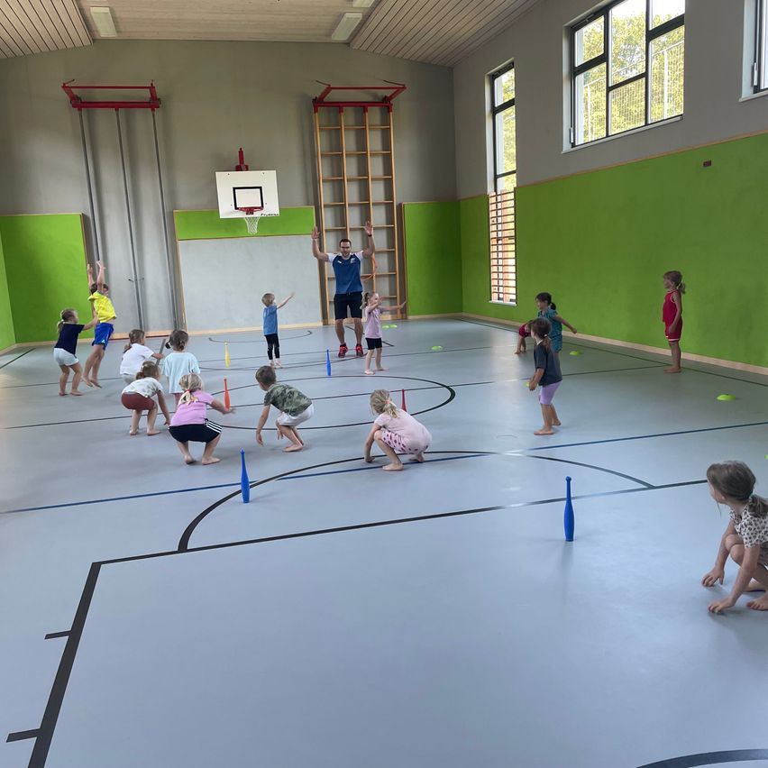 Eine Gruppe von Kindern und ein erwachsener Mann spielen ein Spiel auf einem Spielfeld, mit verstreuten Kegeln und Bällen. Der Mann steht in der Mitte und die Kinder sind in verschiedene Aktivitäten verwickelt.