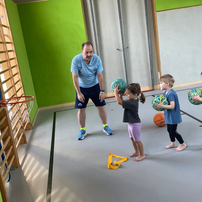 Ein Mann gibt zwei Kindern in einer grün gestrichenen Halle Anweisungen zum Werfen eines Balls. Die Kinder haben bunte Bälle und sind barfuß.