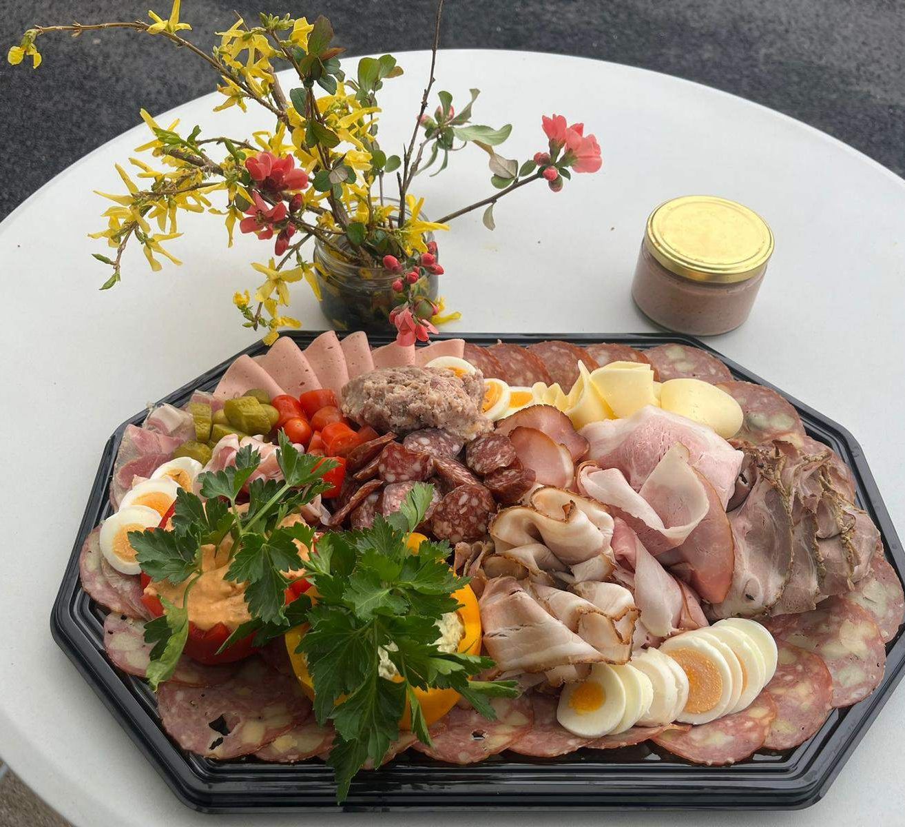 Ein Tablett mit verschiedenen Fleischsorten, Käse und Gemüse, mit Blumen garniert, auf einem weißen Tisch platziert.