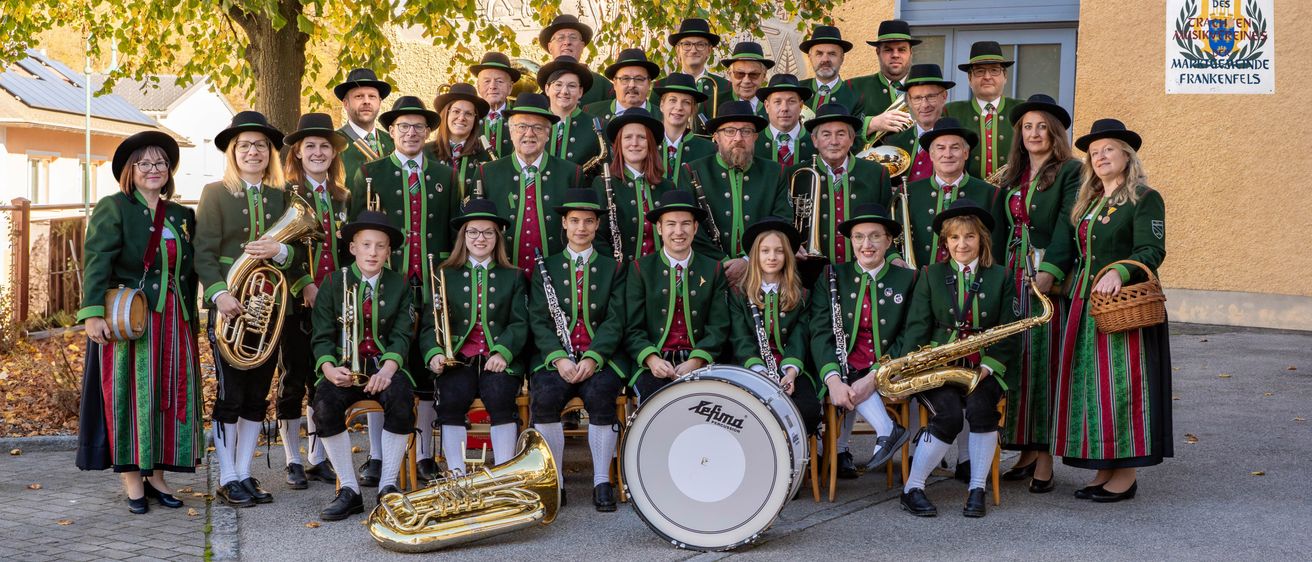 Eine Musikkapelle posiert für ein Gruppenfoto, gekleidet in grünen und roten Uniformen. Zu den Instrumenten gehören Trompeten, Posaunen und eine Trommel. Der Ort ist im Freien in der Nähe eines Gebäudes mit einem Baum im Hintergrund.