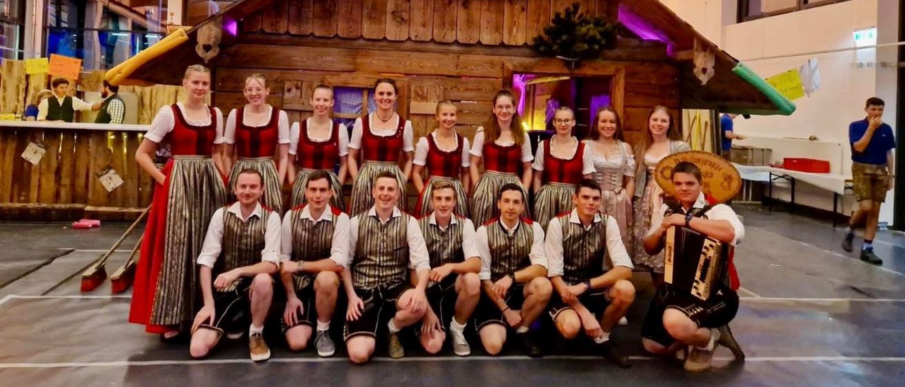 Eine Gruppe von Menschen in traditioneller Kleidung posiert vor einem dekorierten Holzhaus mit einer roten Blumenarrangement.