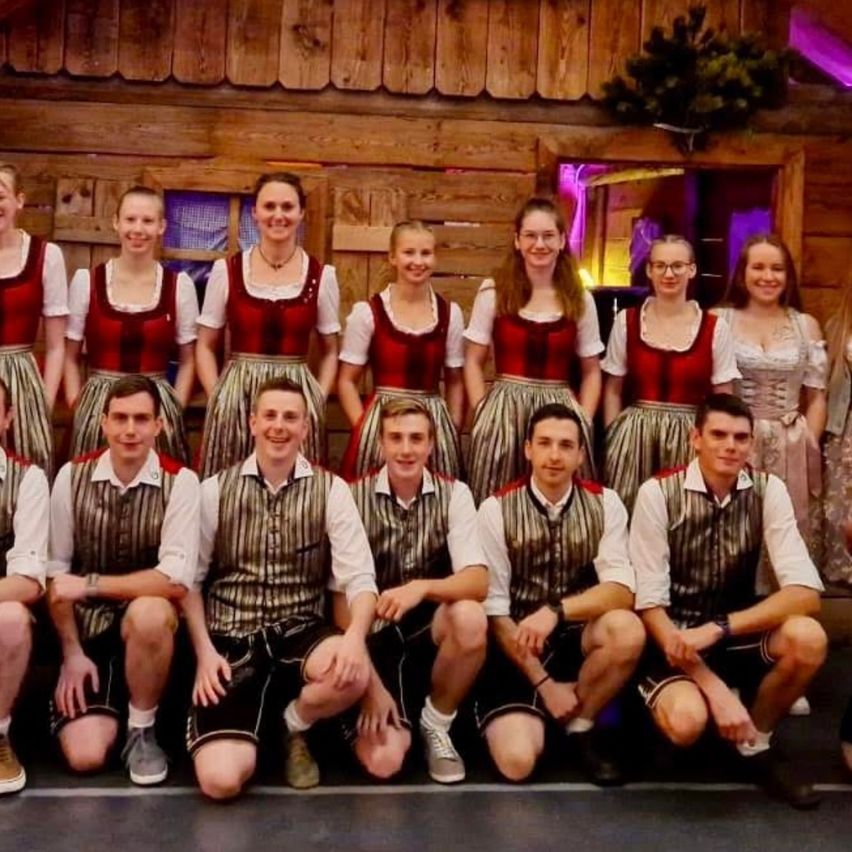 Eine Gruppe von Menschen in traditioneller Kleidung posiert vor einem dekorierten Holzhaus mit einer roten Blumenarrangement.