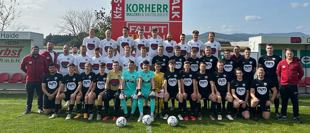 Eine Fußballmannschaft in schwarzen und weißen Trikots posiert für ein Foto auf einem Feld mit zwei Fußballbällen. Die Spieler stehen in einer Reihe, zwei sitzend und einer kniend. Dahinter steht ein Schild mit dem Wort 'Korherr' und Zahlen.