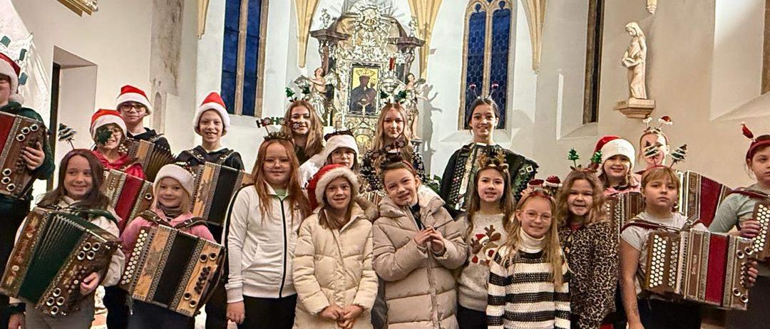Eine Gruppe von Kindern in Weihnachtsmützen und Winterkleidung, die Akkordeons halten und für ein Foto in einer Kirche posieren.