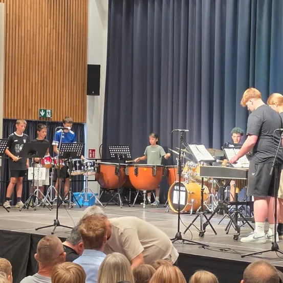 Eine Gruppe junger Musiker spielt auf der Bühne mit verschiedenen Instrumenten, darunter Trommeln und Xylophon, während ein Publikum zuschaut.