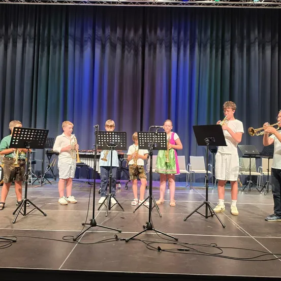 Eine Gruppe von Kindern in Sommerkleidung spielt auf einer Bühne mit Musikinstrumenten. Sie stehen vor Notenständern und einem Hintergrund.
