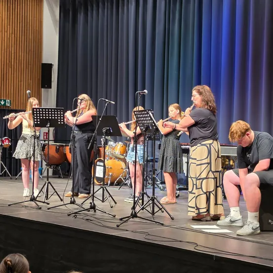 Eine Gruppe von Frauen und einem Mann spielt auf der Bühne Flöten und Trommeln. Die Bühne ist gut beleuchtet, und der Hintergrund besteht aus blauen Vorhängen.