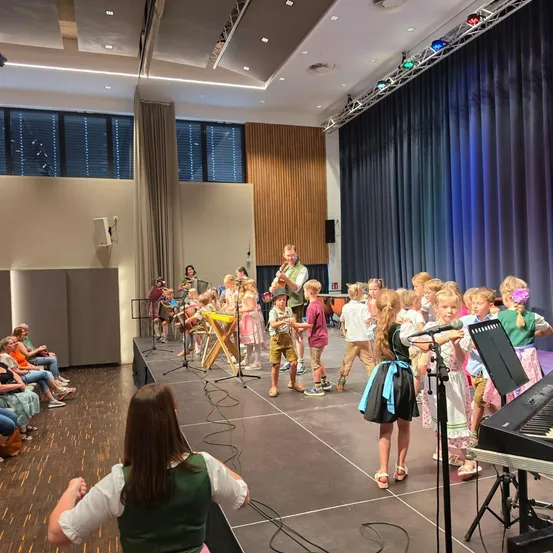 Eine Gruppe von Kindern und Erwachsenen tritt mit Musikinstrumenten auf der Bühne auf, während ein Mann sie anführt. Das Publikum sitzt im Hintergrund.