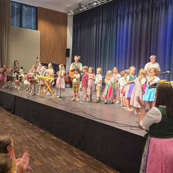 Eine Gruppe von Kindern in traditioneller deutscher Kleidung tritt mit Musikinstrumenten auf der Bühne auf, begleitet von einem Erwachsenen. Die Bühne ist mit einem Hintergrund aus blauen Vorhängen ausgestattet.
