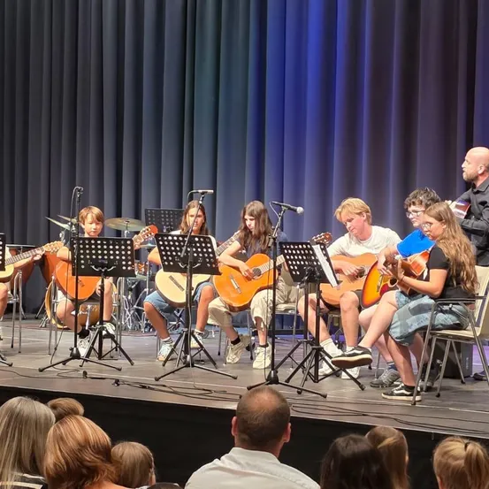 Eine Gruppe junger Musiker, darunter Jungen und Mädchen, tritt mit Gitarren und Trommeln auf der Bühne auf. Sie sitzen auf Stühlen und werden von einem Mann begleitet. Das Publikum beobachtet sie von vorne.