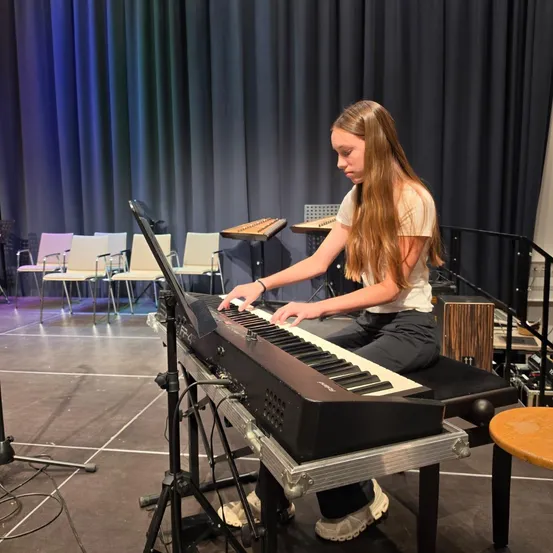 Ein junges Mädchen mit langen Haaren spielt Klavier auf einer Bühne mit einem schwarzen Vorhang im Hintergrund.