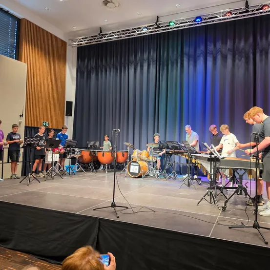 Eine Gruppe junger Musiker spielt auf der Bühne mit verschiedenen Perkussionsinstrumenten unter einem blauen Vorhang.