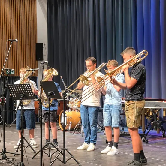 Fünf junge Jungen stehen auf der Bühne und spielen Posaunen. Sie stehen vor Mikrofonen und Notenständern. Hinter ihnen steht ein Schlagzeug und eine Becken.