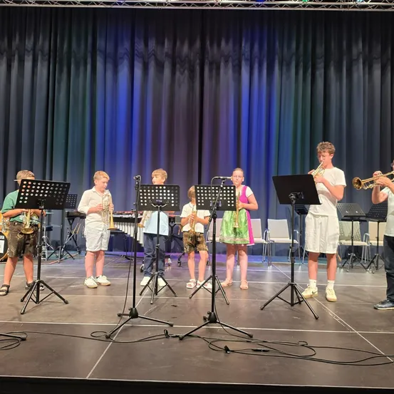 Eine Gruppe von Kindern in weißen und grünen Outfits spielt Musikinstrumente auf der Bühne, mit einem schwarzen Vorhang im Hintergrund.