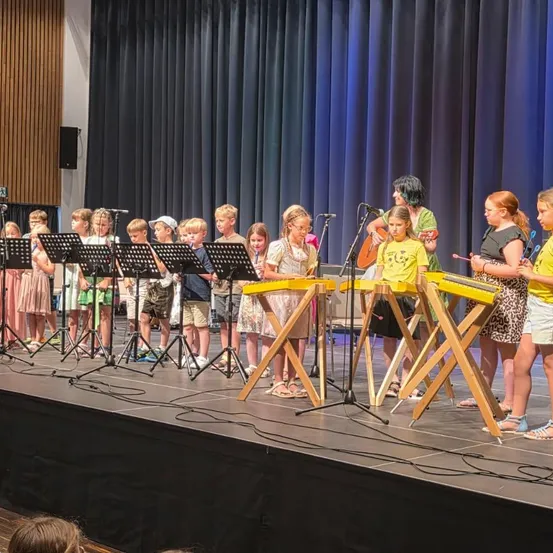 Eine Gruppe von Kindern in gelben Shirts und Shorts spielt auf der Bühne mit Musikinstrumenten, darunter eine Gitarre und ein Xylophon. Eine Frau steht hinter ihnen, möglicherweise ihre Lehrerin.
