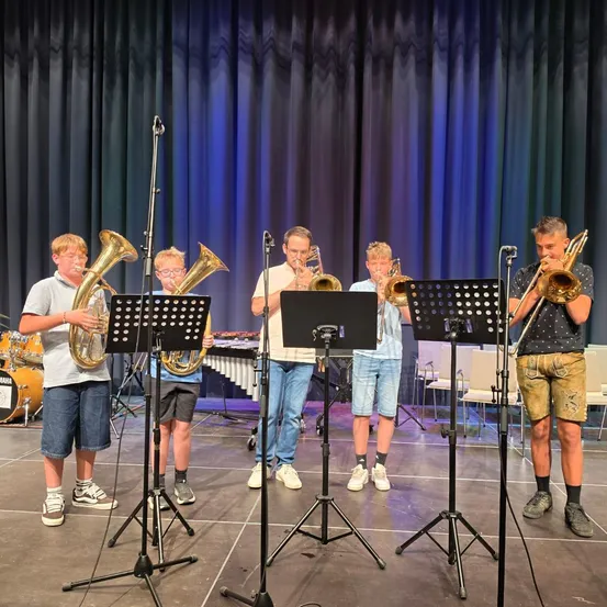 Fünf junge Musiker spielen auf der Bühne Posaunen, vor ihnen Musikstände und Mikrofone. Hinter ihnen ist ein blauer Vorhang und ein Schlagzeug zu sehen.