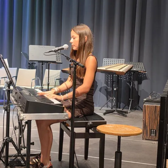 Ein junges Mädchen spielt auf der Bühne ein Keyboard, mit einem Mikrofon vor ihr und verschiedenen Musikinstrumenten im Hintergrund.