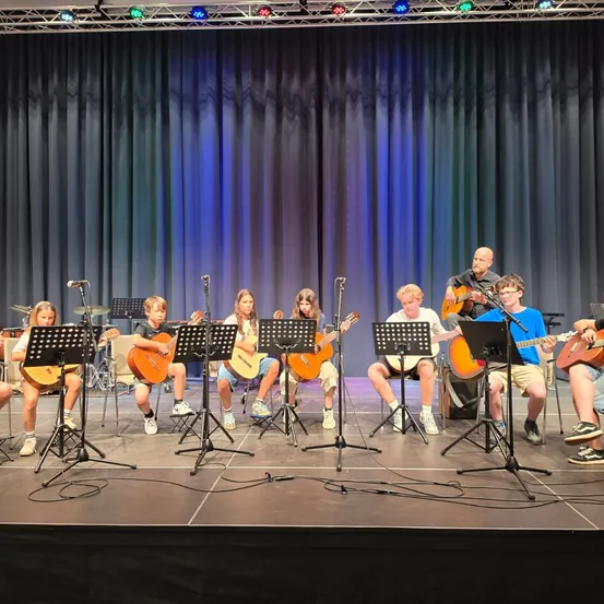 Eine Gruppe von Kindern und zwei Erwachsenen spielt Gitarren auf einer Bühne mit blauer Beleuchtung und einem schwarzen Vorhang im Hintergrund.