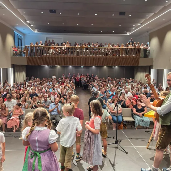 Ein großes Publikum in einem Saal, klatscht und sieht sich eine Aufführung auf der Bühne an. Ein Mann spielt Gitarre und einige Kinder stehen vor ihm.