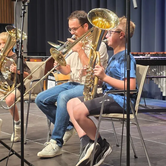 Drei junge Musiker, zwei Jungen und ein erwachsener Mann, sitzen auf der Bühne und spielen jeweils ein Posaune. Der Erwachsene trägt eine Brille und ein weißes Hemd, während die Jungen Freizeitkleidung tragen. Die Bühne ist mit Musikinstrumenten und einem Hintergrund ausgestattet.