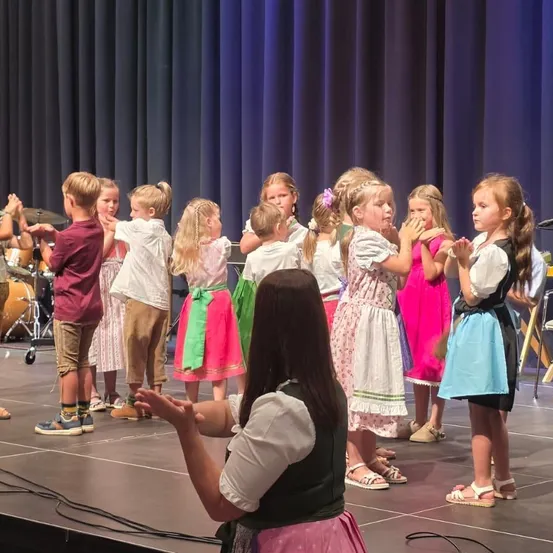 Eine Gruppe von Kindern in traditionellen Kostümen tritt auf der Bühne auf, angeführt von einer Frau. Dahinter steht ein Schlagzeug.