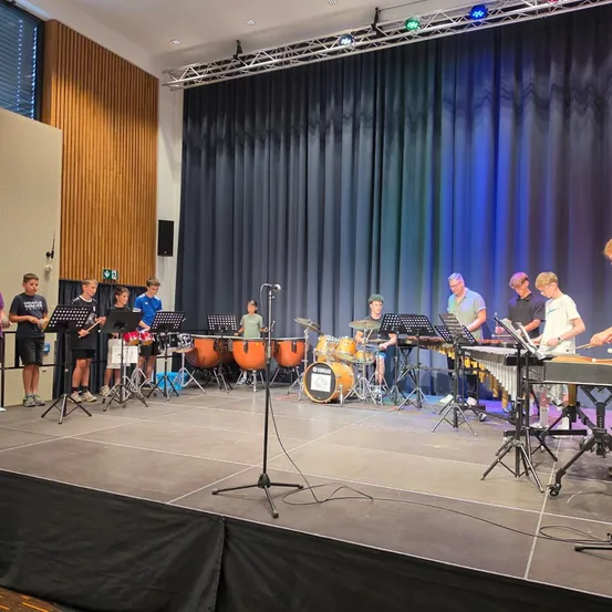 Eine Gruppe junger Musiker spielt auf der Bühne verschiedene Percussionsinstrumente, darunter Trommeln und Xylophon, unter einem blauen Vorhang.