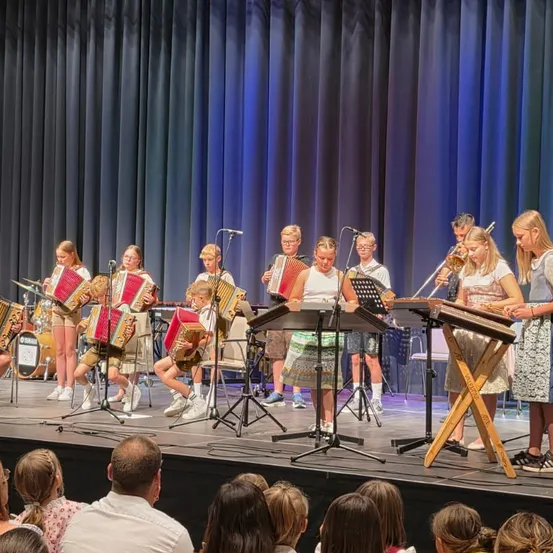 Eine Gruppe von Kindern und Erwachsenen spielt auf der Bühne mit verschiedenen Musikinstrumenten, darunter Akkordeons und Trompeten, vor einem Publikum.