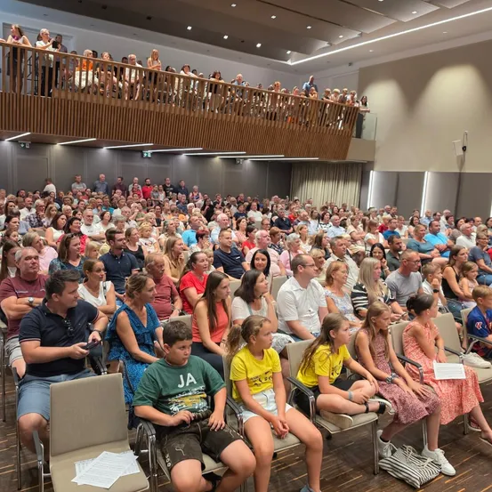 Eine große Menschenmenge sitzt in einem modernen Auditorium, einige stehen auf der Galerie. Das Publikum scheint an einer Veranstaltung teilzunehmen, mit einer Mischung aus Erwachsenen und Kindern.