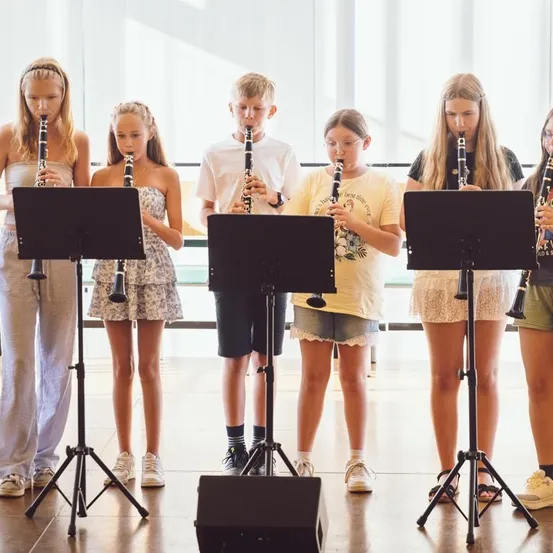 Eine Gruppe junger Musiker, lässig gekleidet, steht in einer Reihe und spielt Klarinetten. Sie stehen vor Notenständern mit schwarzen Rahmen. Der Ort scheint ein Musikraum oder Übungsraum zu sein.