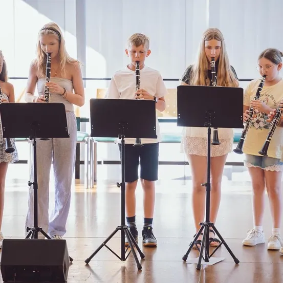Fünf Kinder stehen in einer Reihe und halten jeweils eine Klarinette in der Hand. Sie spielen Musik. Sie befinden sich in einem Raum mit Holzboden und weißen Wänden.