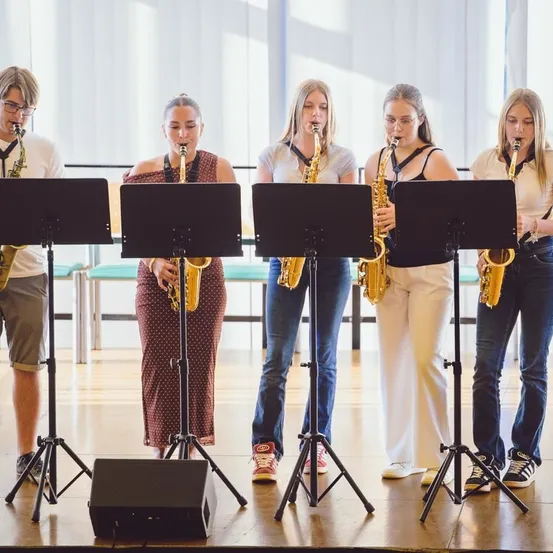 Fünf Personen, vier Frauen und ein Mann, stehen auf einer Bühne und halten jeweils ein Saxophon, das sie spielen. Sie tragen alle Freizeitkleidung und stehen vor Notenständern. Die Bühne hat einen Holzboden und einen weißen Vorhang im Hintergrund.