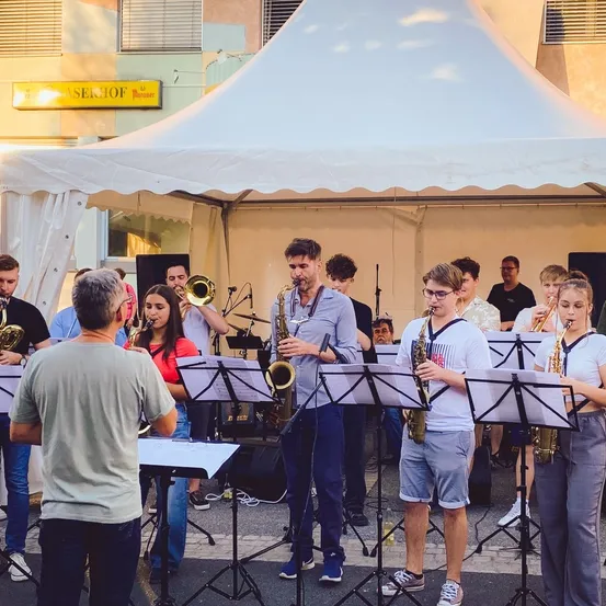 Eine Gruppe von Musikern spielt unter einem Zelt, mit Saxophonen, Trompeten und anderen Instrumenten. Sie sind lässig gekleidet und nehmen wahrscheinlich an einer Straßenaufführung teil.