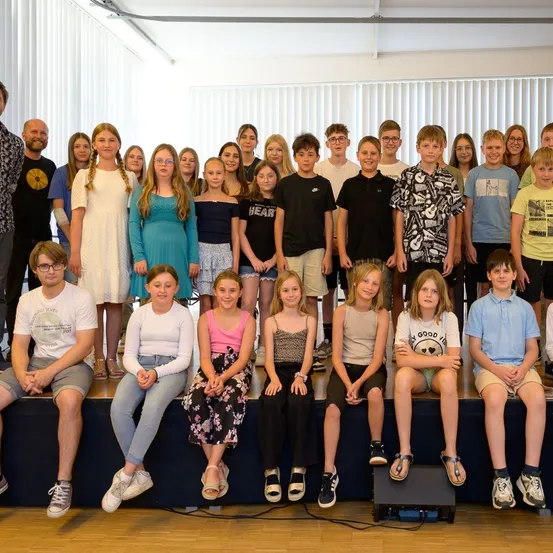 Eine Gruppe junger Menschen, Jungen und Mädchen, posiert für ein Foto in einem Raum mit weißen Jalousien. Sie sitzen und stehen auf einer Bühne.