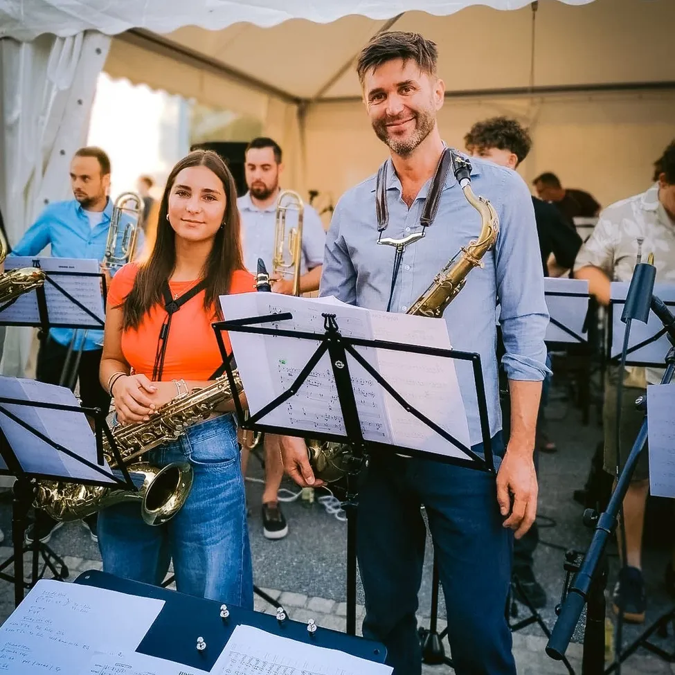 Ein Mann und eine Frau stehen vor einem Notenständer und halten Saxophon. Sie sind von anderen Musikern umgeben, die sich möglicherweise auf eine Aufführung vorbereiten.