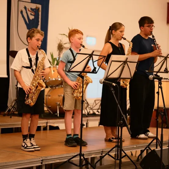 Vier Kinder stehen auf der Bühne und spielen Musikinstrumente. Zwei von ihnen spielen Saxophon, eines spielt Klarinette und das letzte spielt Schlagzeug.