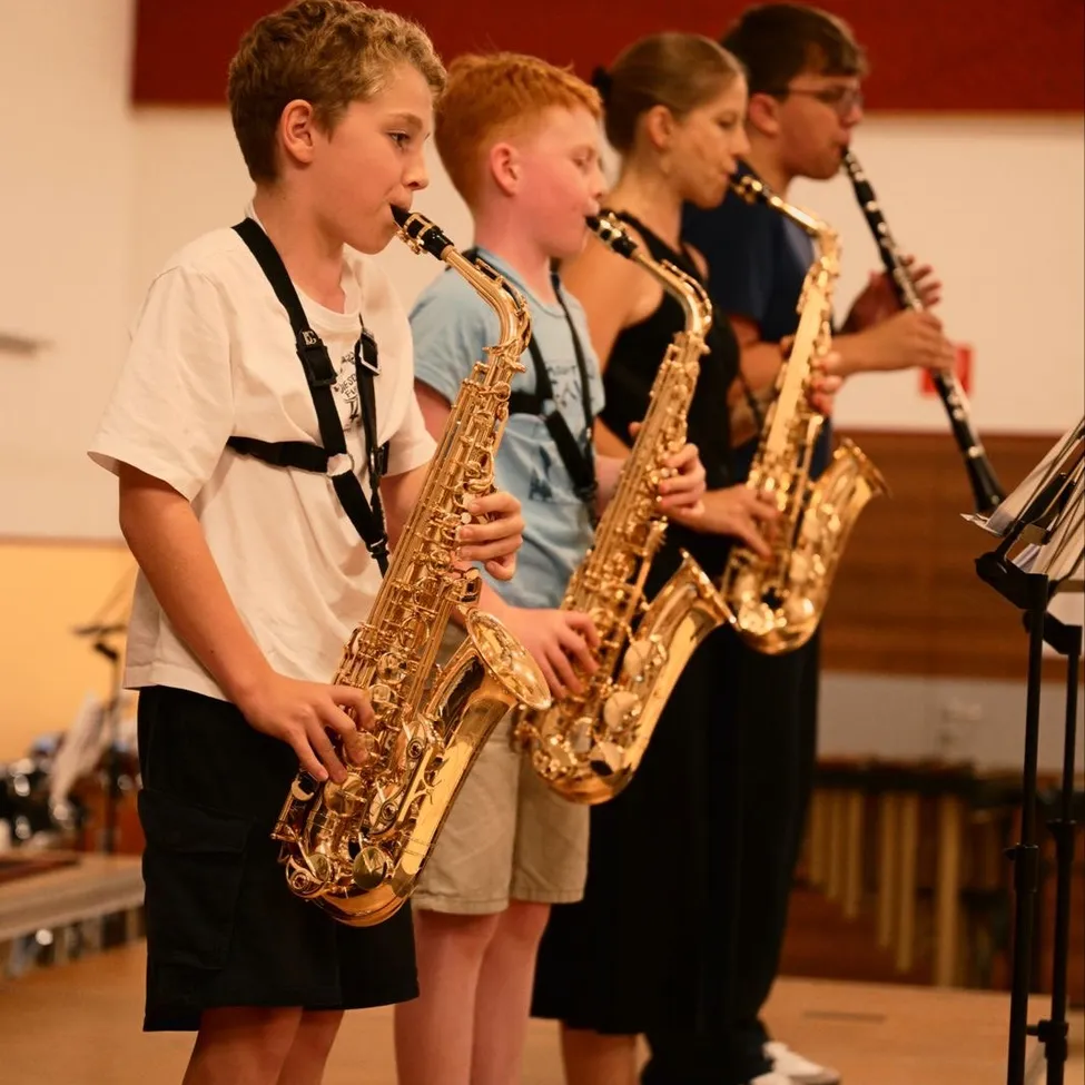 Vier Kinder stehen auf der Bühne und spielen Musikinstrumente. Der Junge auf der linken Seite spielt das Saxophon, und der Junge neben ihm spielt Klarinette. Die beiden Mädchen spielen Saxophon.